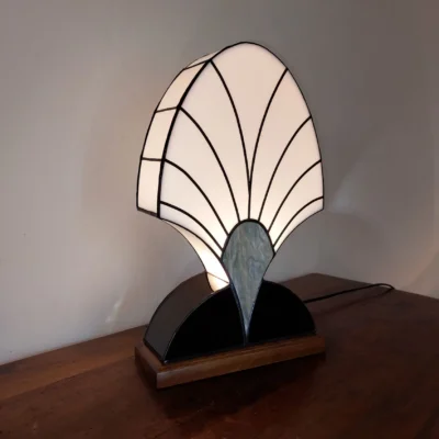 Lampe de table Art Déco en vitrail Tiffany, de forme haute et symétrique Palmette. L'abat-jour est en verre Blanc Opalescent avec des lignes radiales noires. La base est en verre Noir brillant, séparée de l'abat-jour par un motif central triangulaire en verre marbré Gris. Elle repose sur un socle en bois foncé et diffuse une lumière douce mettant en évidence le détail marbré.