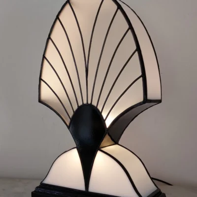 Lampe de table Art Déco en vitrail Tiffany, de forme haute et symétrique, rappelant une palmette stylisée. L'abat-jour est composé de deux sections incurvées en verre blanc opalescent séparées par un col en verre noir opaque. La section supérieure présente un motif de lignes radiales noires rappelant un éventail ou des nervures de coquillage. La lampe repose sur une base rectangulaire noire et diffuse une lumière douce, mettant en valeur son élégant contraste noir et blanc.