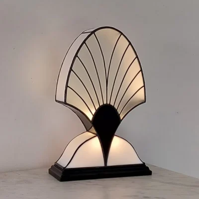 Lampe de table Art Déco en vitrail Tiffany, de forme haute et symétrique, rappelant une palmette stylisée. L'abat-jour est composé de deux sections incurvées en verre blanc opalescent séparées par un col en verre noir opaque. La section supérieure présente un motif de lignes radiales noires rappelant un éventail ou des nervures de coquillage. La lampe repose sur une base rectangulaire noire et diffuse une lumière douce, mettant en valeur son élégant contraste noir et blanc.