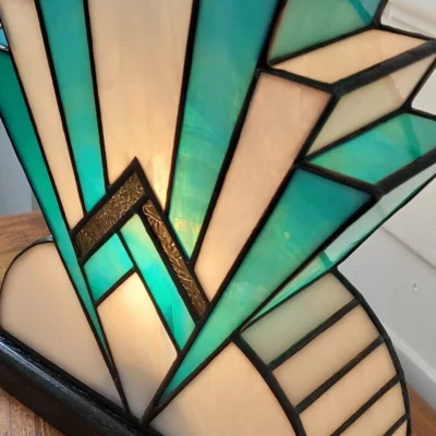 Lampe artisanale en vitrail Tiffany réversible en verre opalescent blanc et bleu canard avec touche de verre or, posée sur un socle en chêne massif sculpté teinté noir, diffusant une lumière chaleureuse.
