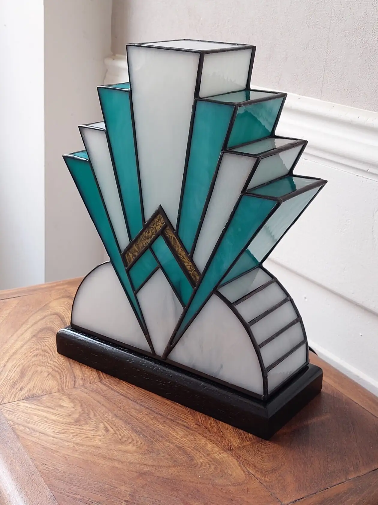 Lampe artisanale en vitrail Tiffany réversible en verre opalescent blanc et bleu canard avec touche de verre or, posée sur un socle en chêne massif sculpté teinté noir, diffusant une lumière chaleureuse.