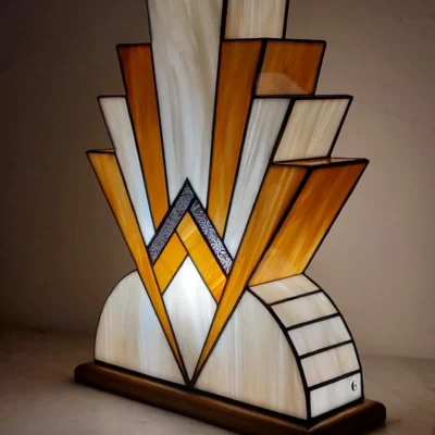 Lampe artisanale en vitrail Art déco avec verres opalescents ivoire et miel caramélisé, motifs géométriques des années 20, posée sur un socle en chêne massif sculpté teinté chêne doré, diffusant une lumière chaleureuse.