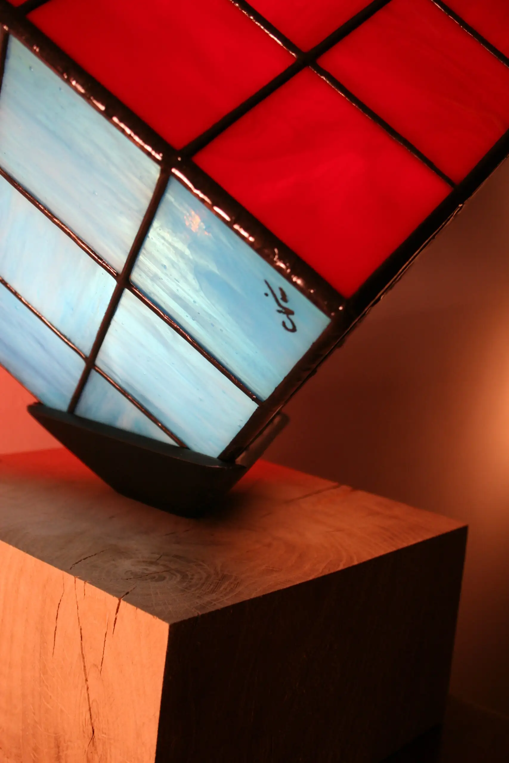 Lampe de table au design de Rubik's Cube, en vitrail coloré. L'abat-jour est un cube en verre serti de lignes de plomb noires épaisses. Trois des faces visibles du cube sont de couleurs différentes : rouge vif, jaune vif, et blanc opalescent avec des nuances de bleu clair. Le cube est posé en équilibre sur un coin, reposant sur un petit support noir. La base de la lampe est un bloc de bois clair. La lumière intérieure illumine intensément les verres colorés, créant une ambiance vive.