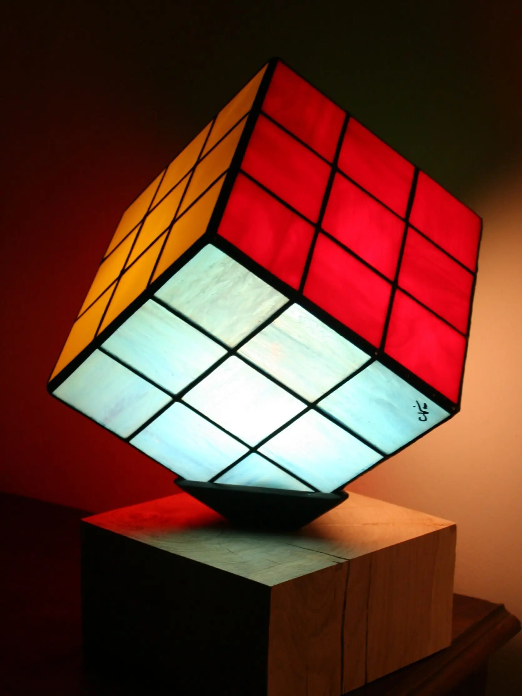 Lampe de table au design de Rubik's Cube, en vitrail coloré. L'abat-jour est un cube en verre serti de lignes de plomb noires épaisses. Trois des faces visibles du cube sont de couleurs différentes : rouge vif, jaune vif, et blanc opalescent avec des nuances de bleu clair. Le cube est posé en équilibre sur un coin, reposant sur un petit support noir. La base de la lampe est un bloc de bois clair. La lumière intérieure illumine intensément les verres colorés, créant une ambiance vive.