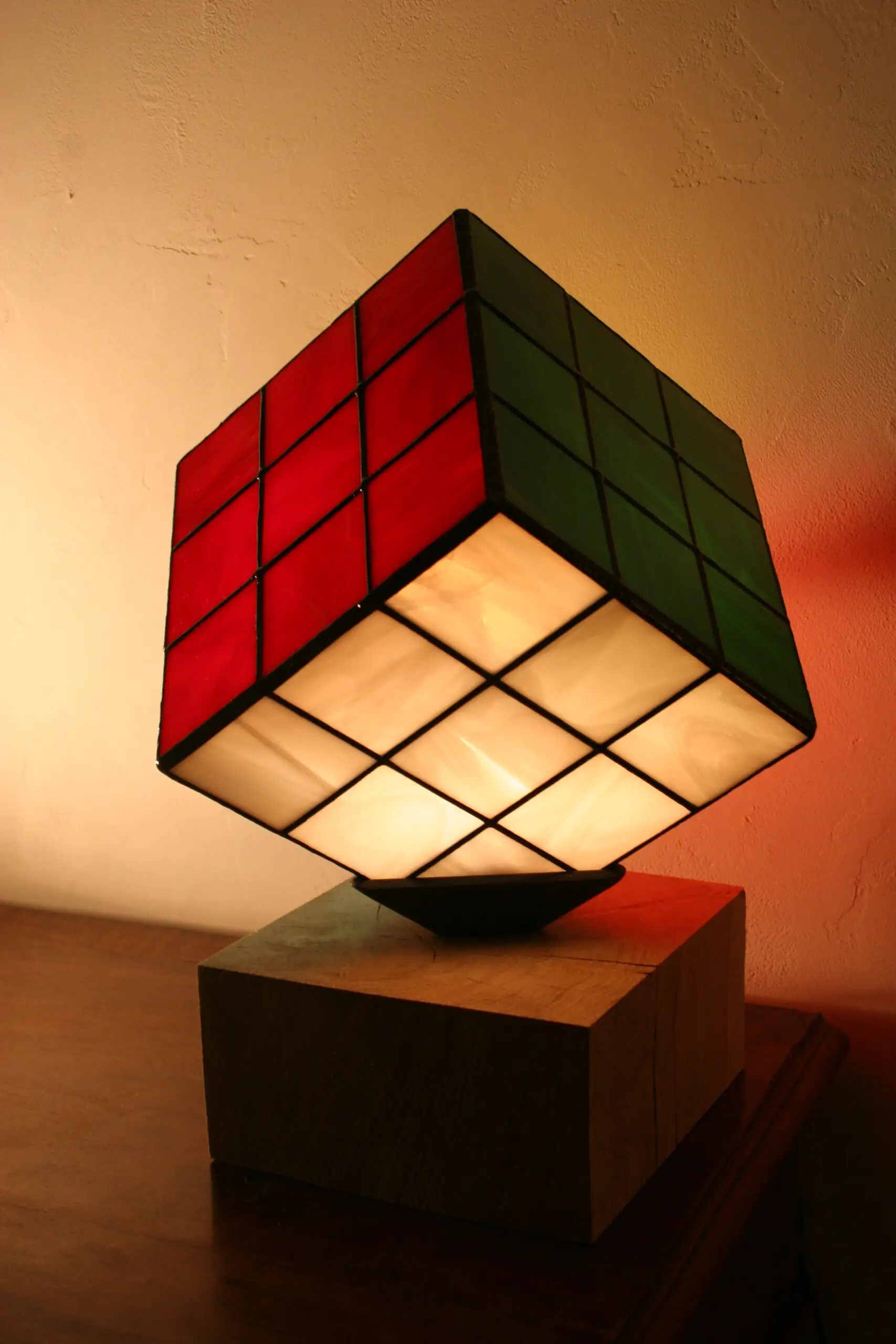 Lampe de table au design de Rubik's Cube, en vitrail coloré. L'abat-jour est un cube en verre serti de lignes de plomb noires épaisses. Trois des faces visibles du cube sont de couleurs différentes : rouge vif, jaune vif, et blanc opalescent avec des nuances de bleu clair. Le cube est posé en équilibre sur un coin, reposant sur un petit support noir. La base de la lampe est un bloc de bois clair. La lumière intérieure illumine intensément les verres colorés, créant une ambiance vive.
