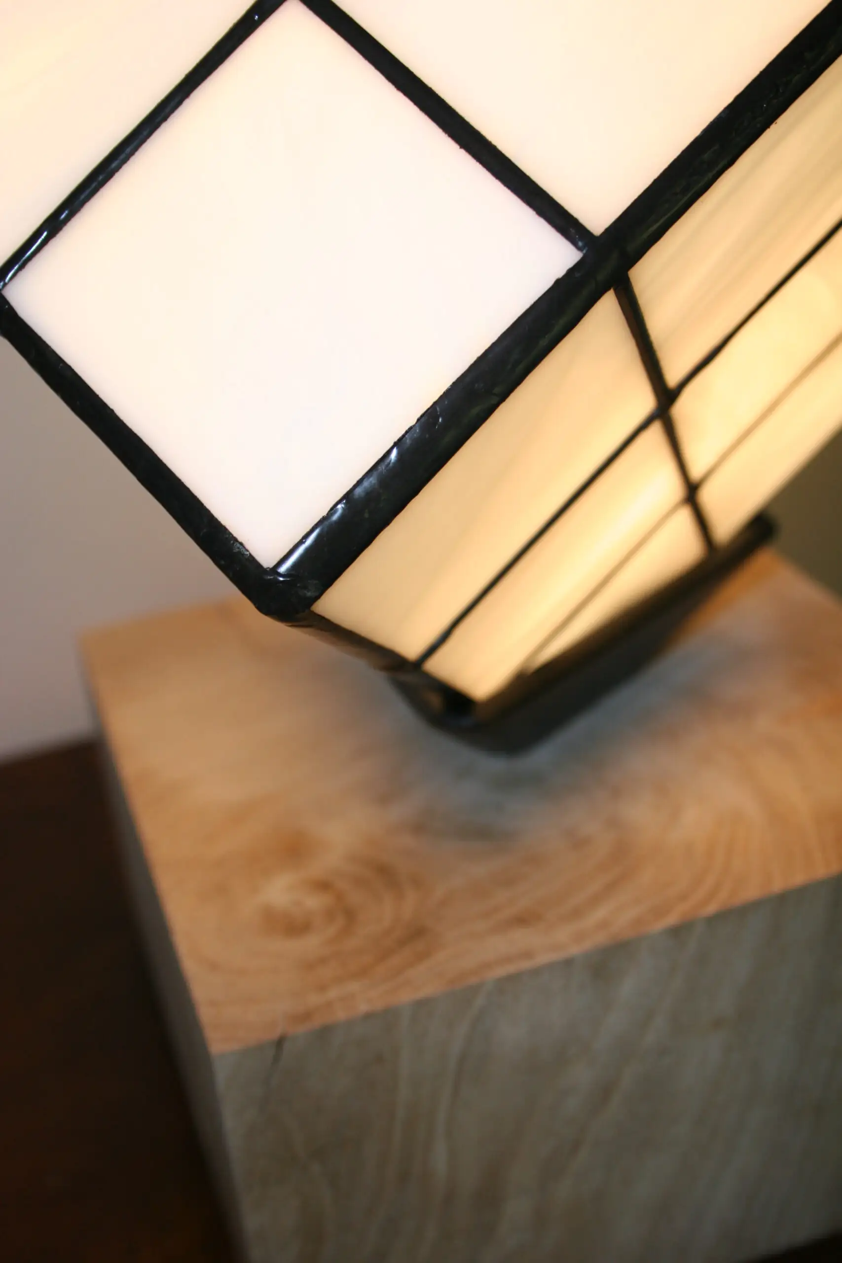 Lampe de table au design unique et ludique, inspirée d'un Rubik's Cube. L'abat-jour est un cube en vitrail de verre blanc opalescent, serti par des lignes de soudures noires qui recréent la grille du célèbre puzzle. Le cube est positionné en équilibre sur un angle, reposant sur un petit support noir. La base de la lampe est un bloc de bois clair, de forme cubique ou parallélépipédique. La lumière douce et chaude émanant du vitrail illumine le verre et la pièce environnante.