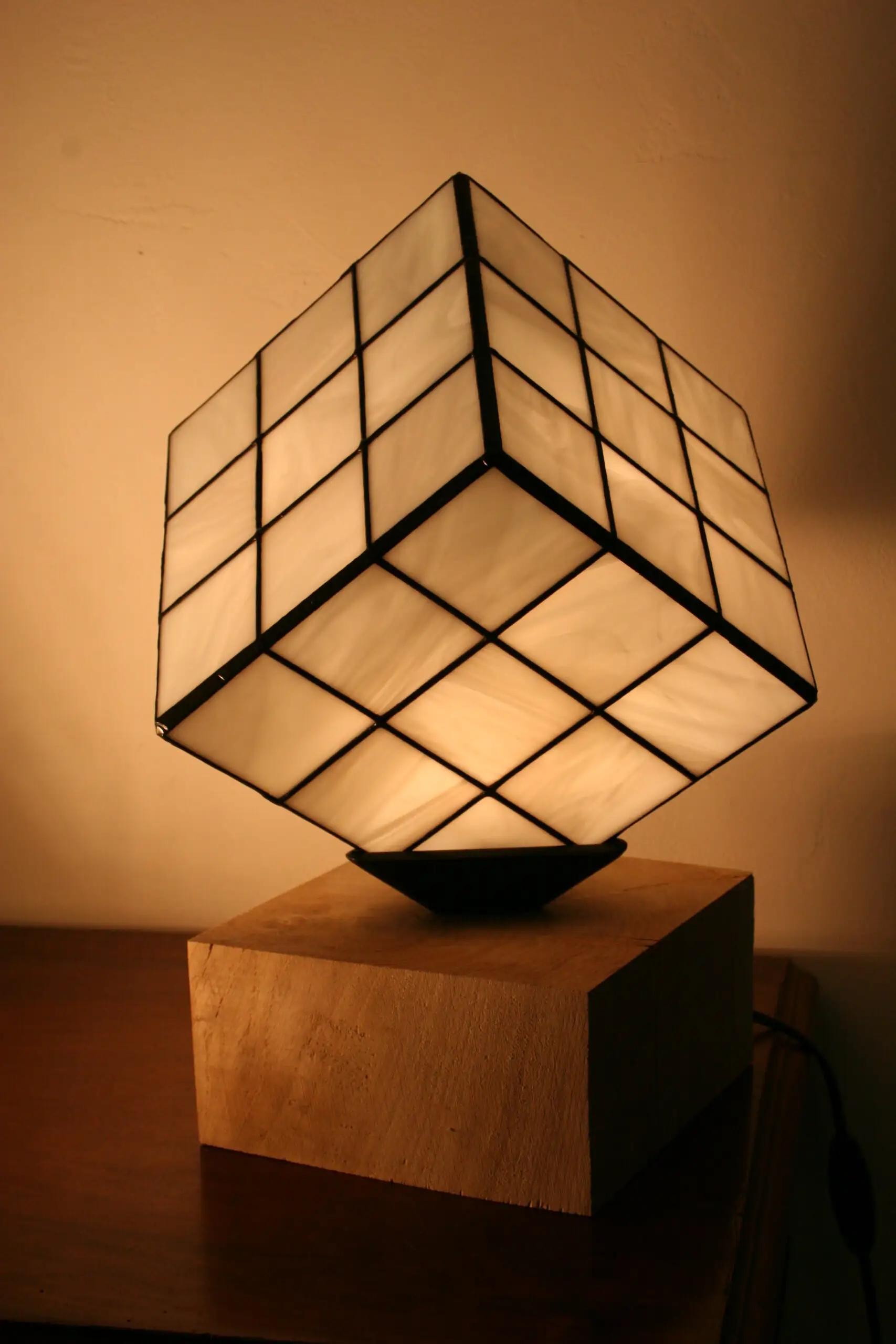 Lampe de table au design unique et ludique, inspirée d'un Rubik's Cube. L'abat-jour est un cube en vitrail de verre blanc opalescent, serti par des lignes de soudures noires qui recréent la grille du célèbre puzzle. Le cube est positionné en équilibre sur un angle, reposant sur un petit support noir. La base de la lampe est un bloc de bois clair, de forme cubique ou parallélépipédique. La lumière douce et chaude émanant du vitrail illumine le verre et la pièce environnante.