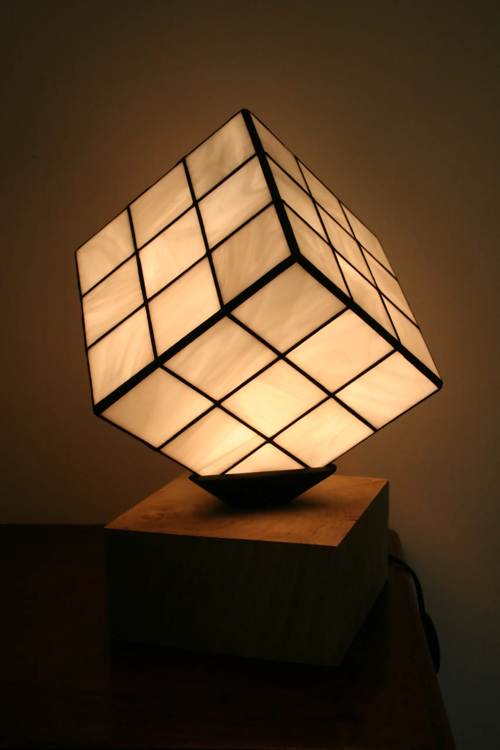 Lampe de table au design unique et ludique, inspirée d'un Rubik's Cube. L'abat-jour est un cube en vitrail de verre blanc opalescent, serti par des lignes de soudures noires qui recréent la grille du célèbre puzzle. Le cube est positionné en équilibre sur un angle, reposant sur un petit support noir. La base de la lampe est un bloc de bois clair, de forme cubique ou parallélépipédique. La lumière douce et chaude émanant du vitrail illumine le verre et la pièce environnante.
