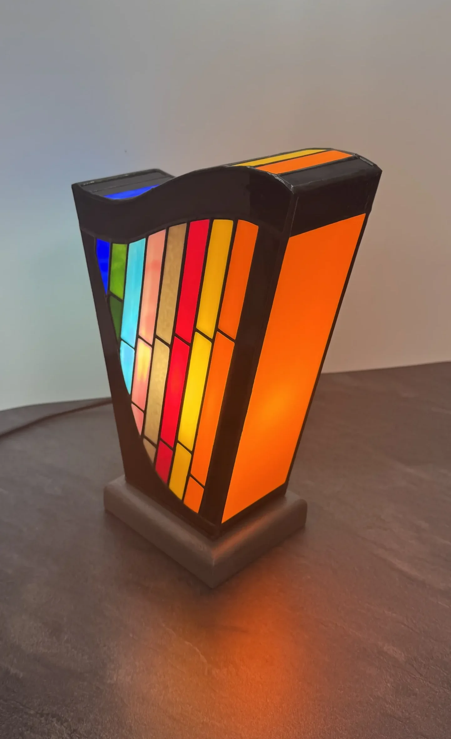 Lampe d’art contemporain Harpège Zénith en vitrail Tiffany, forme de harpe, verres opalescents multicolores sur structure noire et socle en béton ciré.