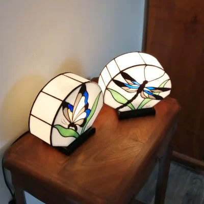 Paire de lampes de table ou de chevets en vitrail, réalisées dans le style Art Nouveau/Tiffany. Les deux lampes ont des abat-jours hémisphériques en verre blanc opalescent, sertis de lignes noires. La lampe de gauche présente un motif de papillon (Butterfly) aux ailes bleues, blanches et marron, entouré de feuilles vertes. La lampe de droite présente un motif de libellule (Dragonfly) aux ailes bleues, jaunes et marron. Les lampes sont posées sur une surface en bois sombre et diffusent une lumière chaude
