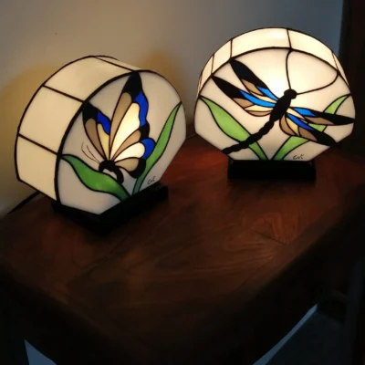 Paire de lampes de table ou de chevets en vitrail, réalisées dans le style Art Nouveau/Tiffany. Les deux lampes ont des abat-jours hémisphériques en verre blanc opalescent, sertis de lignes noires. La lampe de gauche présente un motif de papillon (Butterfly) aux ailes bleues, blanches et marron, entouré de feuilles vertes. La lampe de droite présente un motif de libellule (Dragonfly) aux ailes bleues, jaunes et marron. Les lampes sont posées sur une surface en bois sombre et diffusent une lumière chaude