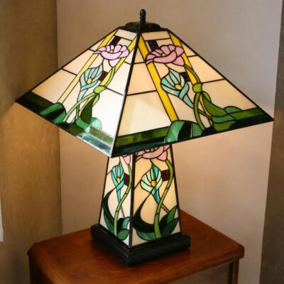 Grande lampe de table ou de bureau en vitrail Tiffany de style Art Nouveau. Elle possède un pied et un abat-jour de forme pyramidale et carrée. L'abat-jour et la base sont décorés de motifs floraux (probablement des arums ou des lys calla) aux teintes pastel de rose, bleu/turquoise, et jaune pâle, sur un fond de verre blanc opalescent. Des bandes de verre vert foncé encadrent la base et les bords de l'abat-jour. La lampe est posée sur une petite table d'appoint en bois avec un pied sculpté, et elle diffuse une lumière chaude