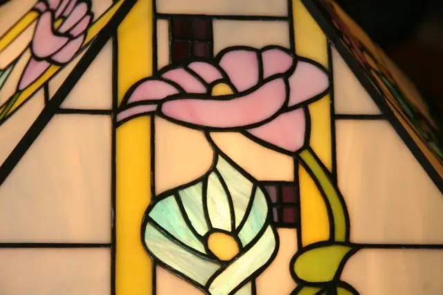 Grande lampe de table ou de bureau en vitrail Tiffany de style Art Nouveau. Elle possède un pied et un abat-jour de forme pyramidale et carrée. L'abat-jour et la base sont décorés de motifs floraux (probablement des arums ou des lys calla) aux teintes pastel de rose, bleu/turquoise, et jaune pâle, sur un fond de verre blanc opalescent. Des bandes de verre vert foncé encadrent la base et les bords de l'abat-jour. La lampe est posée sur une petite table d'appoint en bois avec un pied sculpté, et elle diffuse une lumière chaude