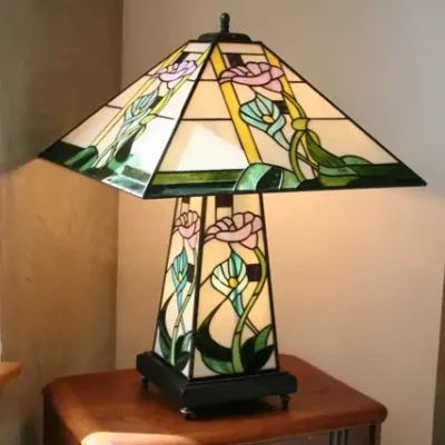 Grande lampe de table ou de bureau en vitrail Tiffany de style Art Nouveau. Elle possède un pied et un abat-jour de forme pyramidale et carrée. L'abat-jour et la base sont décorés de motifs floraux (probablement des arums ou des lys calla) aux teintes pastel de rose, bleu/turquoise, et jaune pâle, sur un fond de verre blanc opalescent. Des bandes de verre vert foncé encadrent la base et les bords de l'abat-jour. La lampe est posée sur une petite table d'appoint en bois avec un pied sculpté, et elle diffuse une lumière chaude