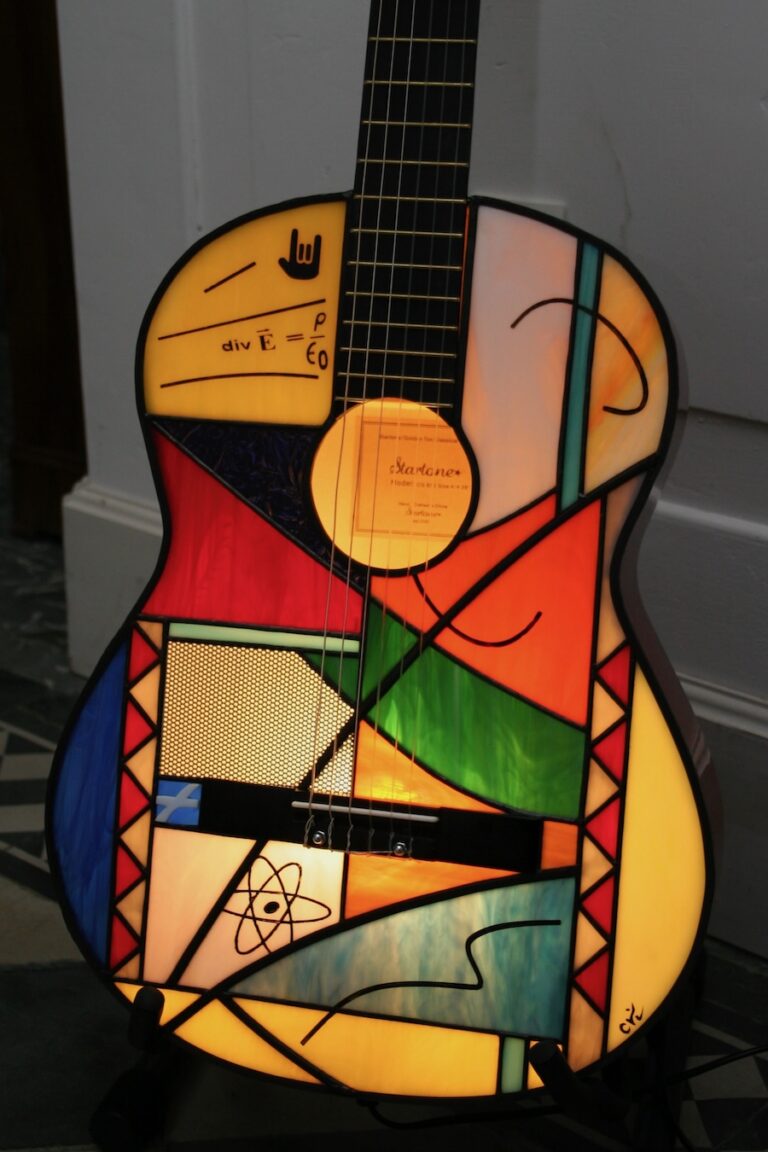 Lampe de sol artistique créée à partir d'une guitare acoustique transformée. La caisse de résonance de la guitare a été remplacée par un panneau en vitrail de style Tiffany, aux motifs abstraits et géométriques. Le vitrail intègre une palette de couleurs vives : bleu ciel, vert émeraude, rouge, orange, jaune, et verre irisé. Le corps en vitrail est éclairé par l'intérieur, illuminant le motif qui inclut de petits détails comme des notes de musique stylisées près de l'ouïe. La guitare est posée sur un support de sol noir sur un carrelage effet bois