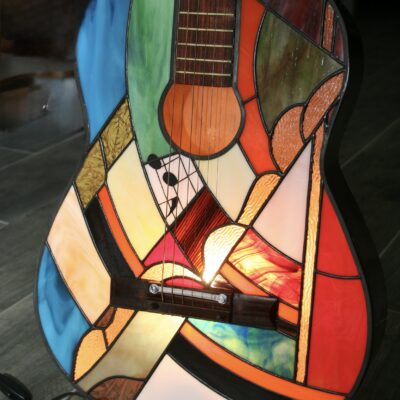 Lampe de sol artistique créée à partir d'une guitare acoustique transformée. La caisse de résonance de la guitare a été remplacée par un panneau en vitrail de style Tiffany, aux motifs abstraits et géométriques. Le vitrail intègre une palette de couleurs vives : bleu ciel, vert émeraude, rouge, orange, jaune, et verre irisé. Le corps en vitrail est éclairé par l'intérieur, illuminant le motif qui inclut de petits détails comme des notes de musique stylisées près de l'ouïe.