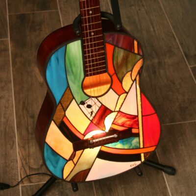 Lampe de sol artistique créée à partir d'une guitare acoustique transformée. La caisse de résonance de la guitare a été remplacée par un panneau en vitrail de style Tiffany, aux motifs abstraits et géométriques. Le vitrail intègre une palette de couleurs vives : bleu ciel, vert émeraude, rouge, orange, jaune, et verre irisé. Le corps en vitrail est éclairé par l'intérieur, illuminant le motif qui inclut de petits détails comme des notes de musique stylisées près de l'ouïe. La guitare est posée sur un support de sol noir sur un carrelage effet bois.