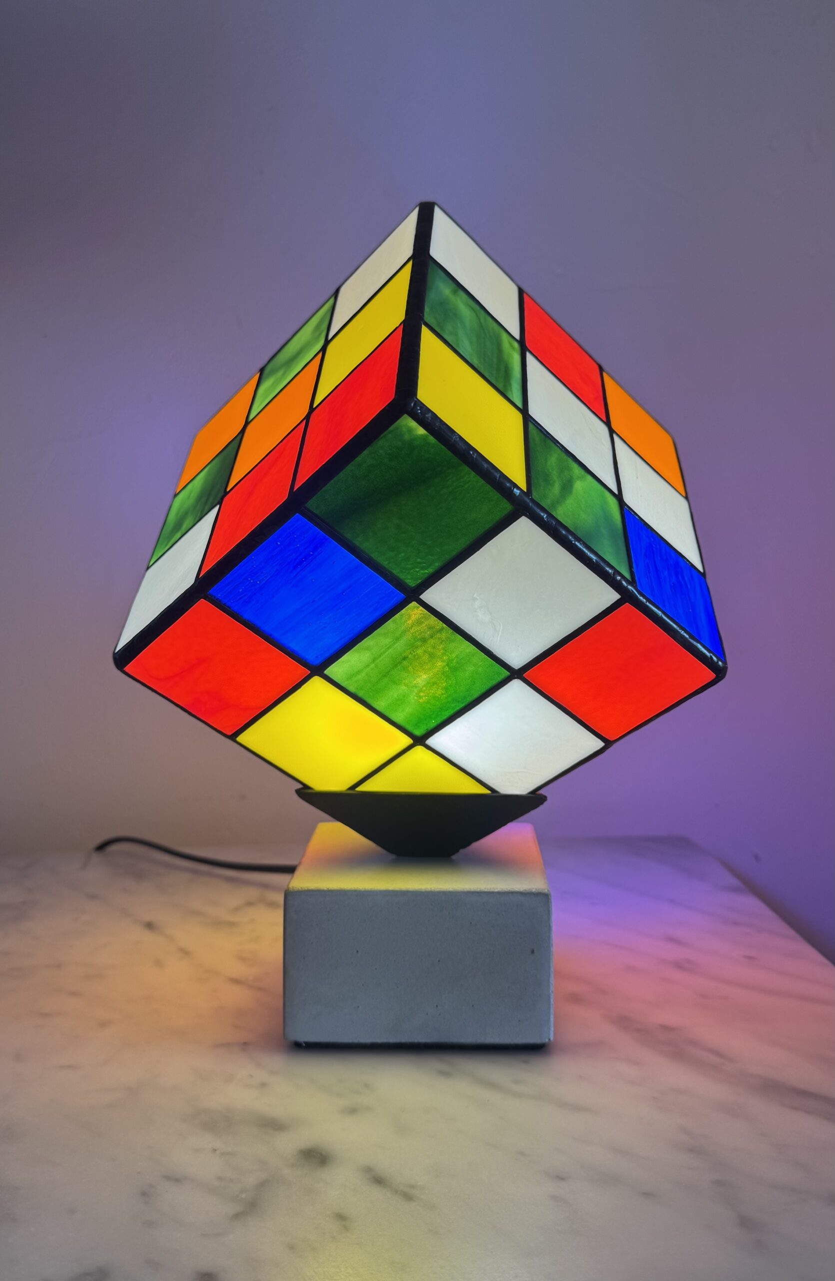 Lampe Rubik's Cube en vitrail Tiffany sur socle béton, luminaire artisanal français multicolore de style design contemporain.