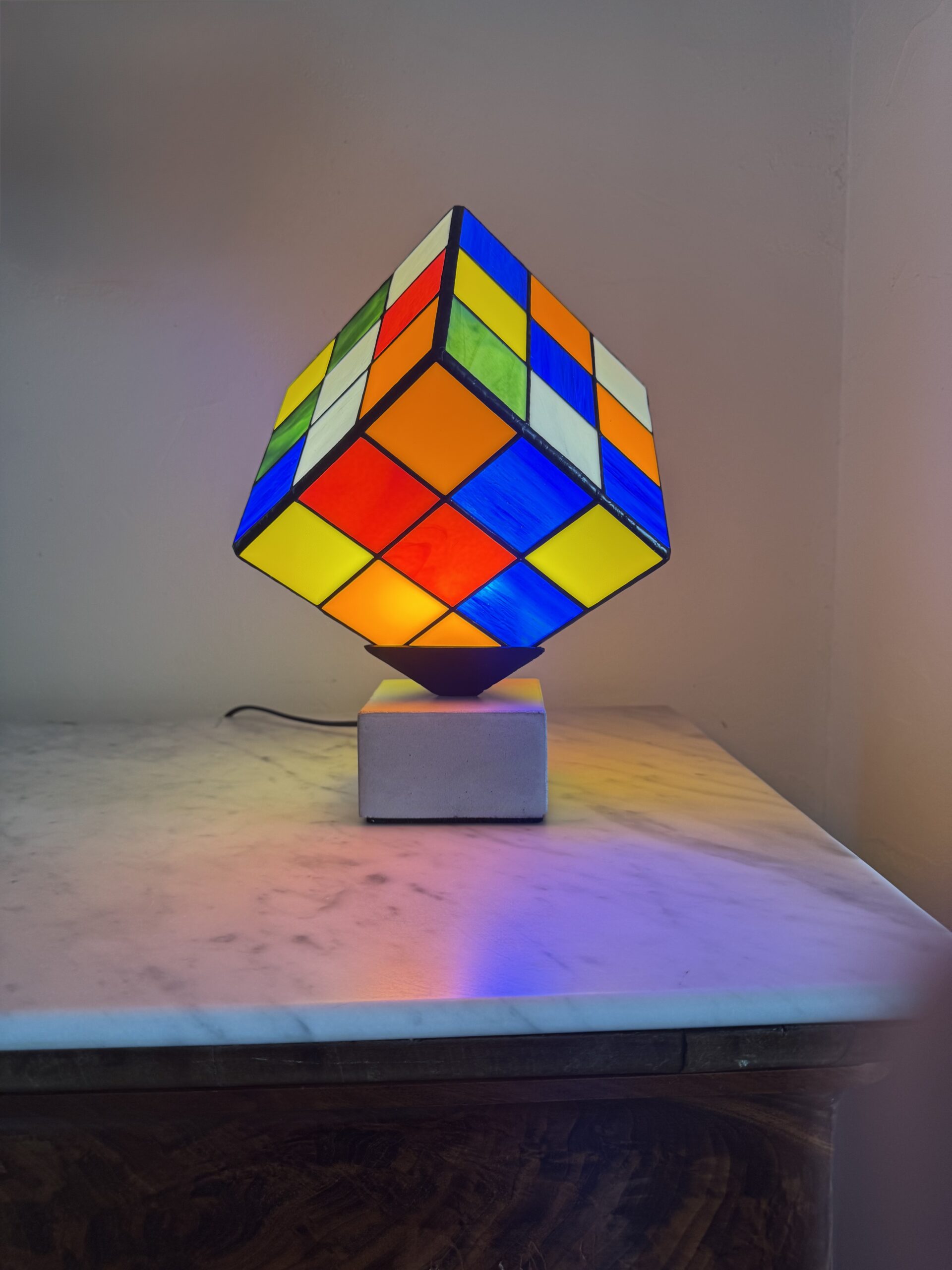 Lampe Rubik's Cube en vitrail Tiffany sur socle béton, luminaire artisanal français multicolore de style design contemporain.